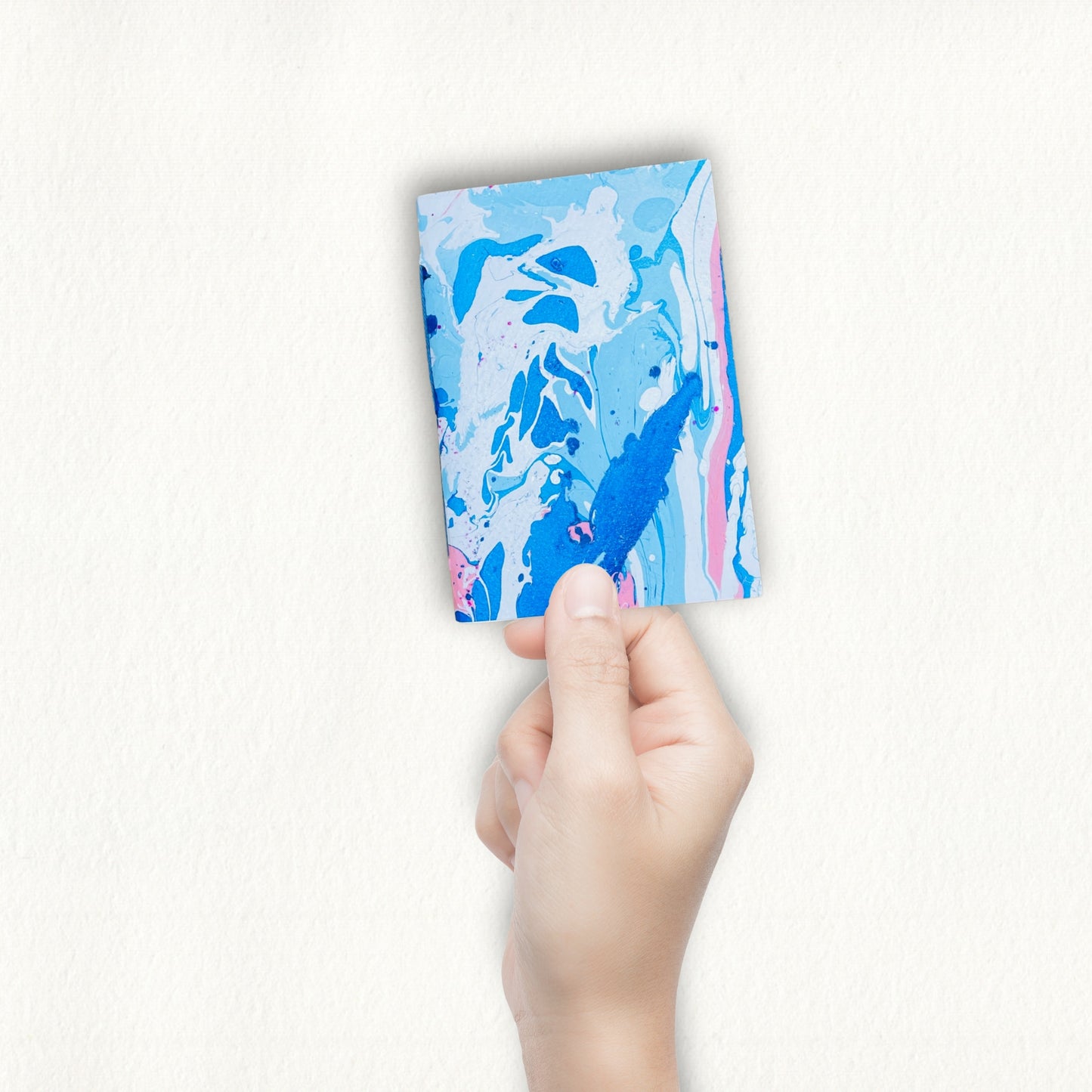Hand hält das Aven Notizbuch mit fließendem blauen und rosa Marmormuster vor schlichtem, weißem Hintergrund.