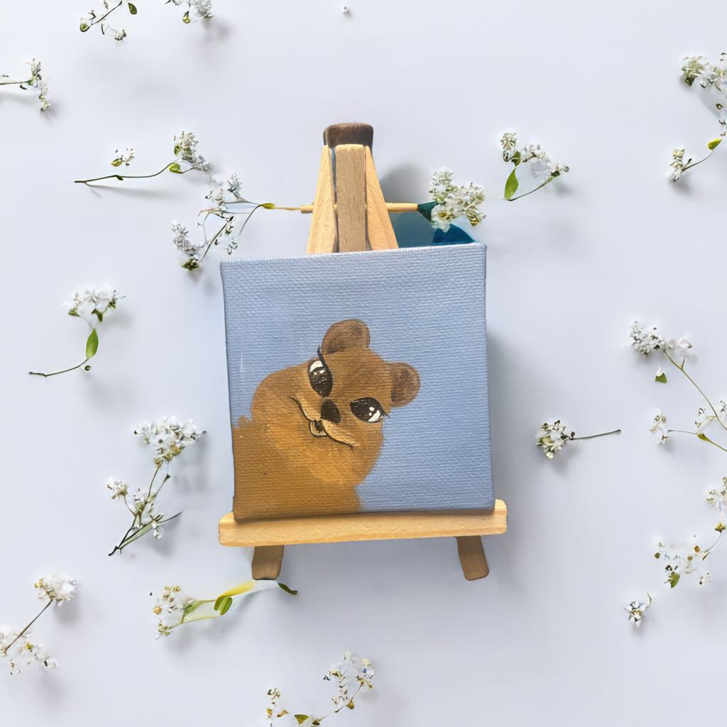 Mini Leinwand 7x7 cm mit handgemaltem Quokka auf hellblauem Hintergrund, auf Holzstaffelei dekoriert mit weißen Blüten – freundliche Mini-Deko.