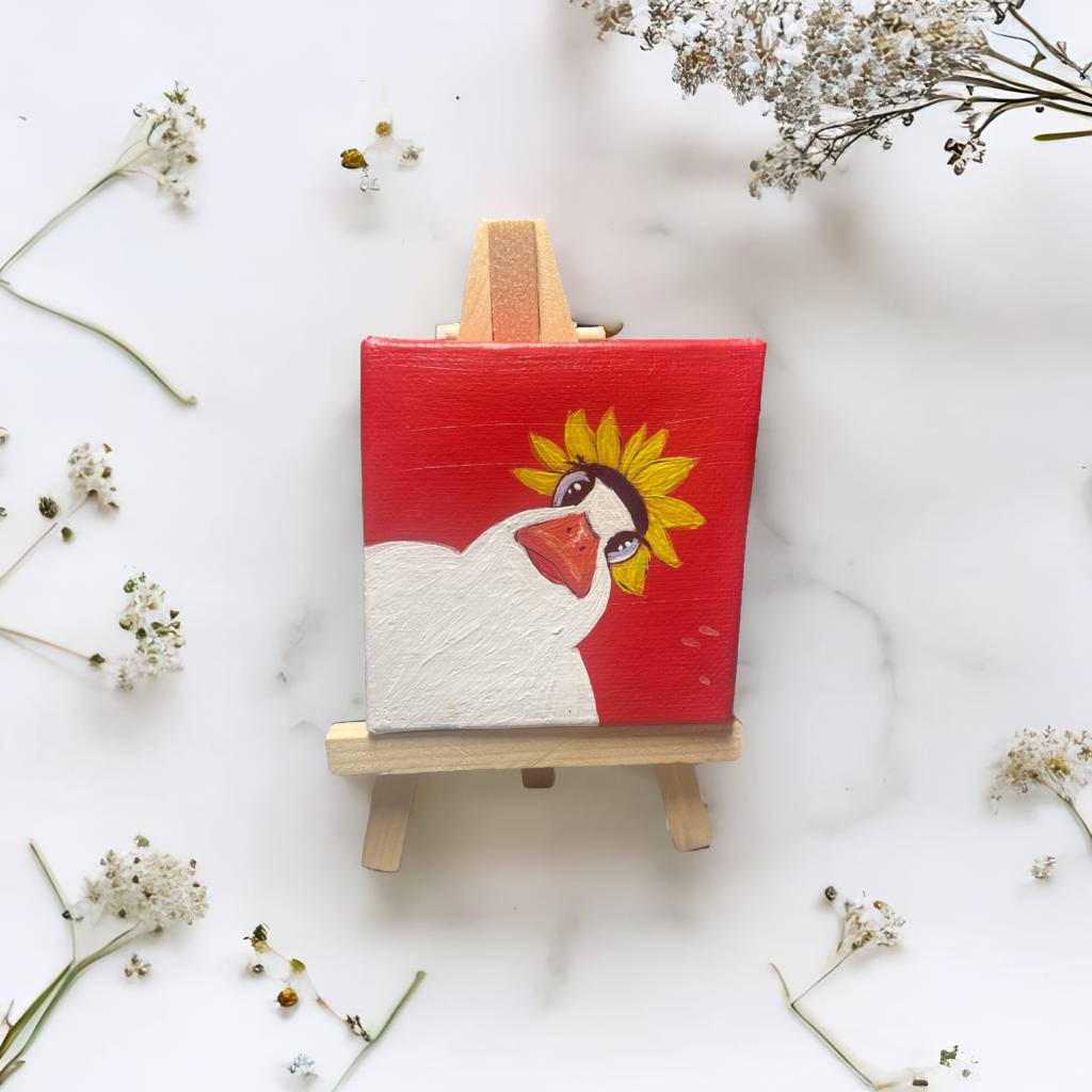 Handgemalte Mini Leinwand 7x7 cm mit Ente im Sonnenblumenhut, auf Holzstaffelei stehend – charmante Deko mit Humor.