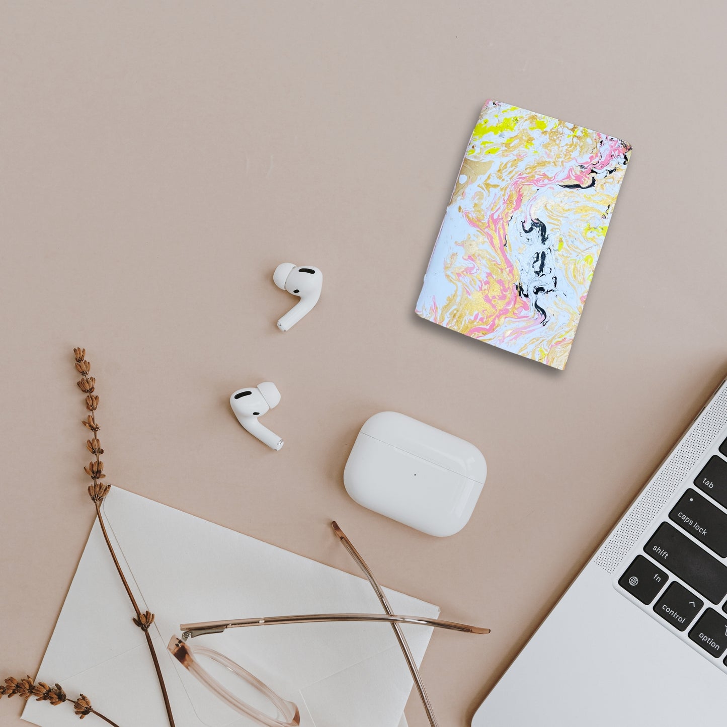 Kreatives Arrangement mit Seluna, AirPods, Brille und Macbook auf beigem Untergrund.