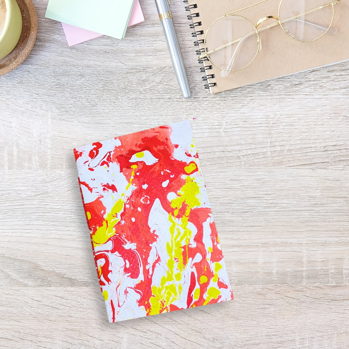 Marmoriertes Notizbuch in Rot-Gelb auf einem Schreibtisch, daneben ein MacBook, Stift und Kaffeetasse.