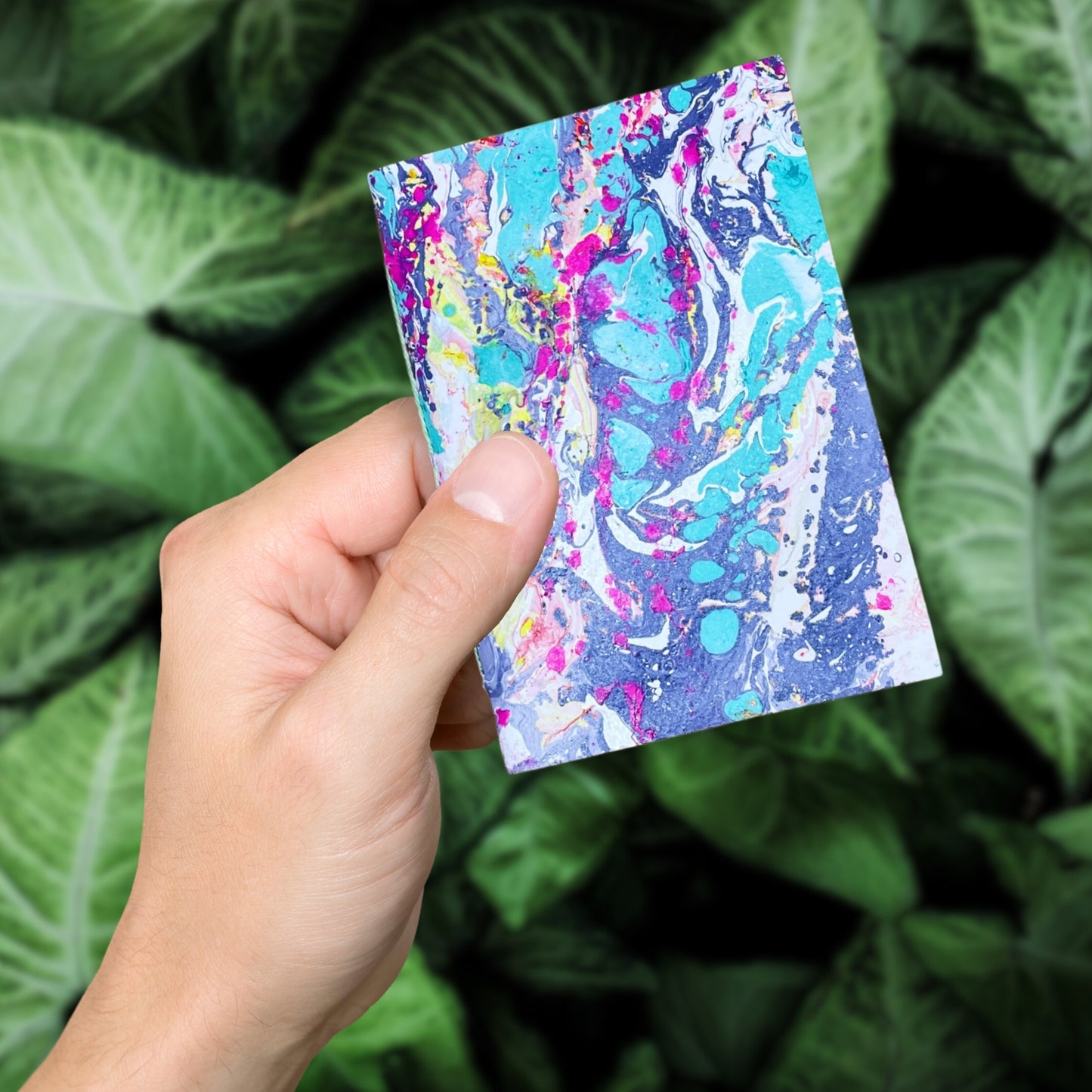 Eine Hand hält das Notizbuch Ysra vor grünen Blättern – das Cover leuchtet intensiv violett.