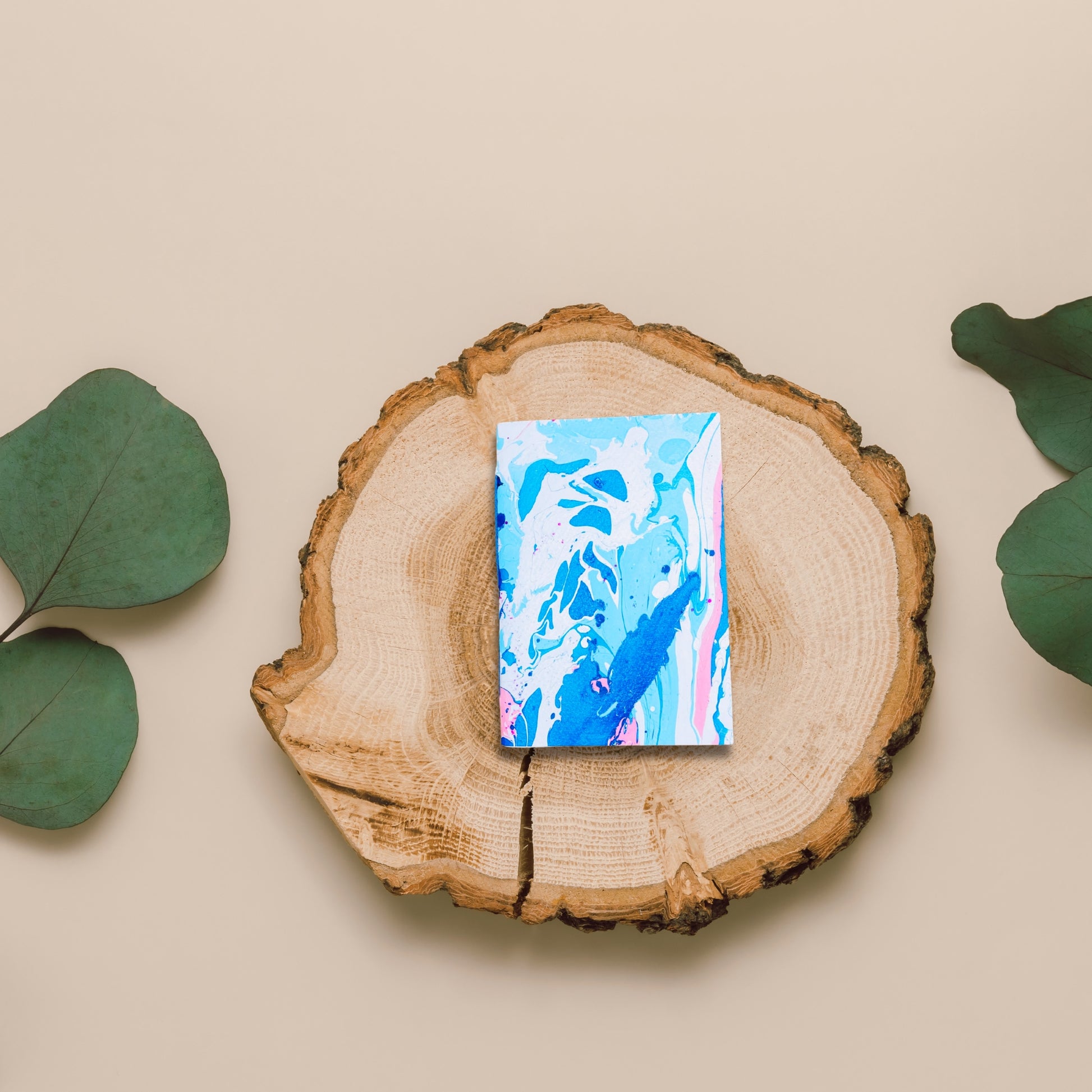 Handgebundenes Notizbuch Aven mit blauer, weißer und pinker Marmormusterung, dekorativ auf einer Holzplatte mit Eukalyptusblättern präsentiert.