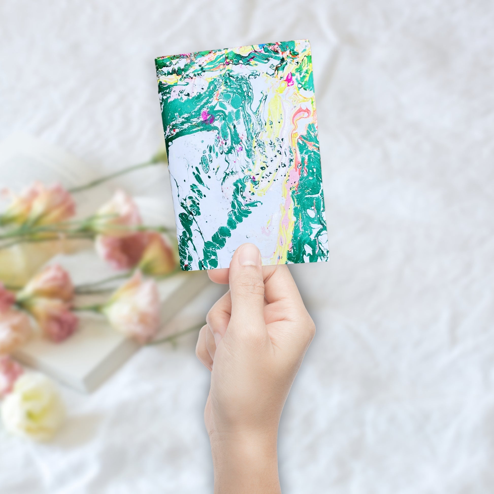 Hand hält das Evora Notizbuch vor unscharfem Hintergrund mit Blumen – perfekt als liebevolles Geschenk oder Tagebuch.