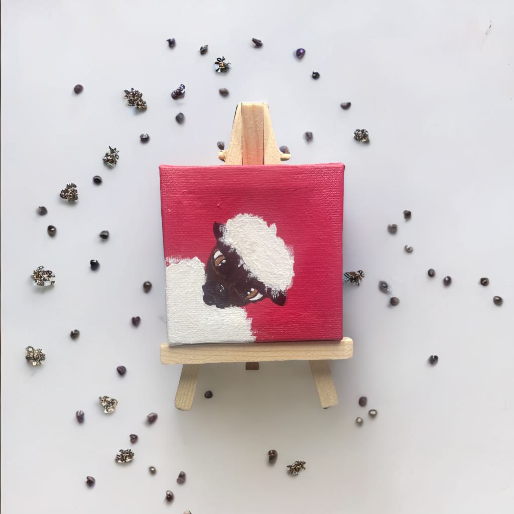 Handgemalte Mini Leinwand 7x7 cm mit schwarzem Schaf auf rotem Hintergrund, auf Holzstaffelei platziert und umgeben von kleinen glänzenden Perlen.

