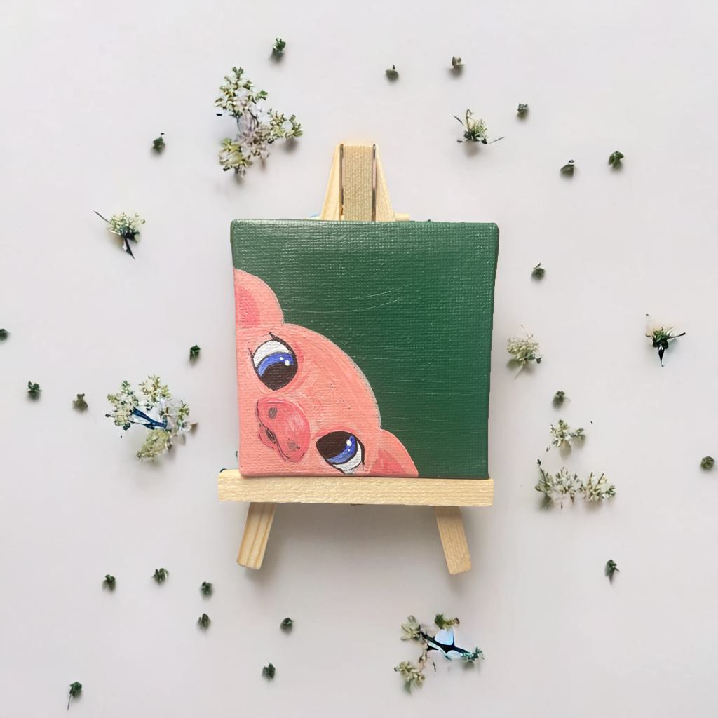 Mini-Leinwand (7×7 cm) mit handgemaltem Schweinchen, platziert auf einer kleinen Holzstaffelei, umgeben von filigranen Blüten – liebevolle Deko oder Geschenkidee.