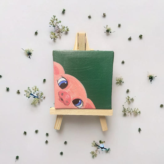 Mini-Leinwand (7×7 cm) mit handgemaltem Schweinchen, platziert auf einer kleinen Holzstaffelei, umgeben von filigranen Blüten – liebevolle Deko oder Geschenkidee.