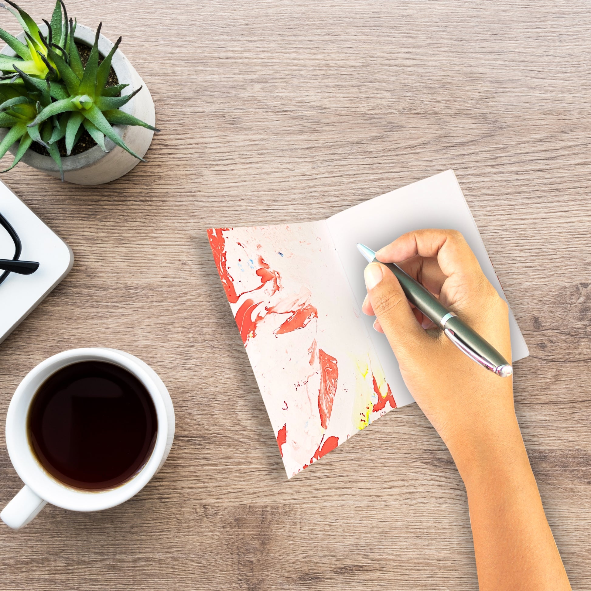 Eine Person schreibt in das geöffnete Notizbuch Oris auf einem Holztisch – Kaffee und Pflanze im Bild.