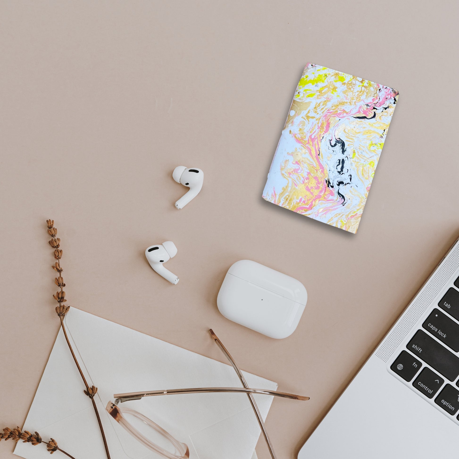 Kreatives Arrangement mit Seluna, AirPods, Brille und Macbook auf beigem Untergrund.