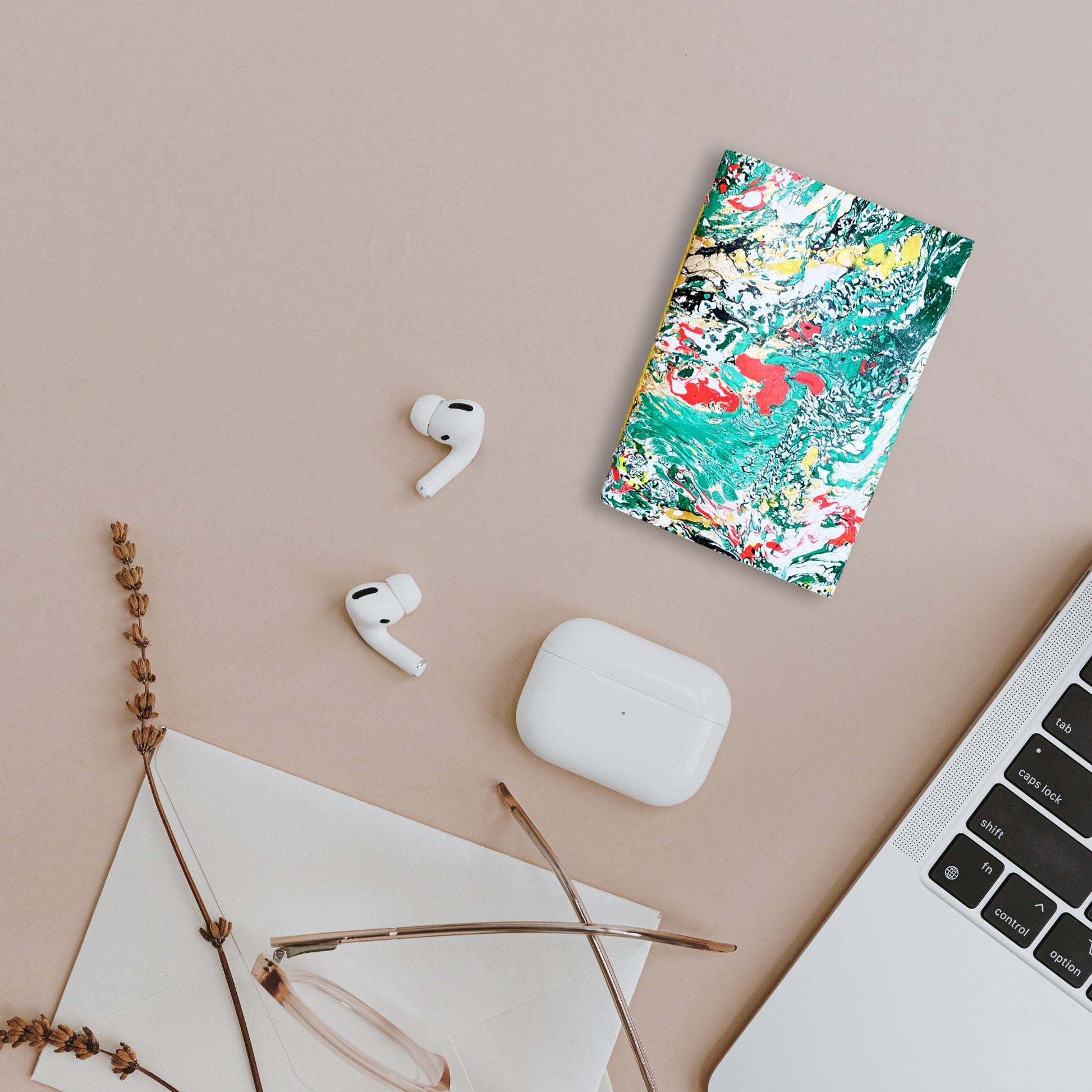 Szene mit Notizbuch Servin, AirPods und MacBook – flach auf beigem Tisch.