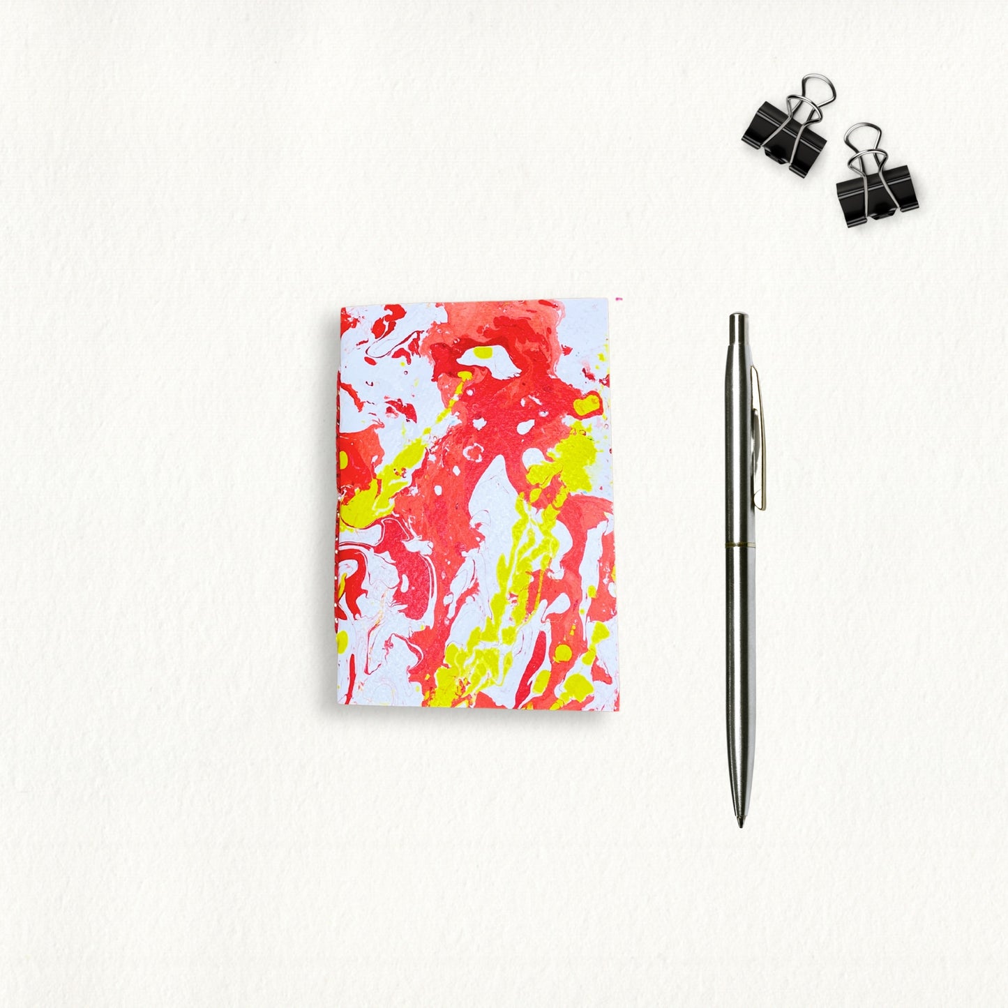 Handgebundenes A6-Notizbuch mit marmoriertem Cover in Rot und Gelb, daneben ein silberner Stift, flach auf beigem Hintergrund fotografiert.